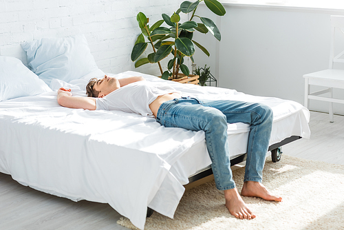 handsome man in t-shirt and jeans lying on back on bed in bedroom
