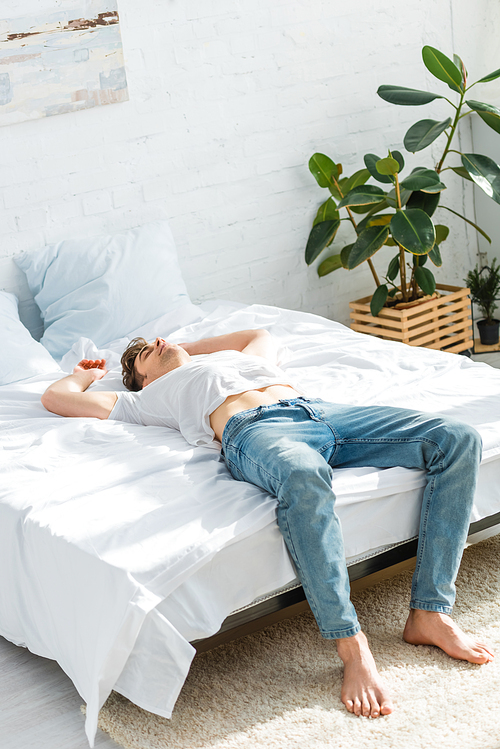 man in t-shirt and jeans lying on back on bed in bedroom