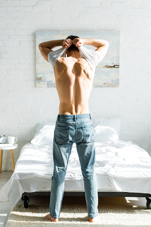 man in jeans standing backwards near bed and taking off white t-shirt in bedroom
