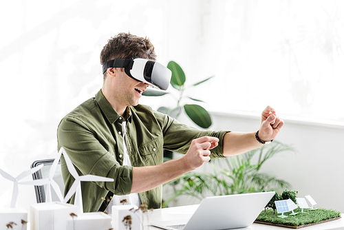architect in reality headset at table with laptop and windmills, buildings, solar panels models in office