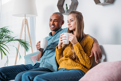 multicultural couple sitting on sofa with coffee and looking away