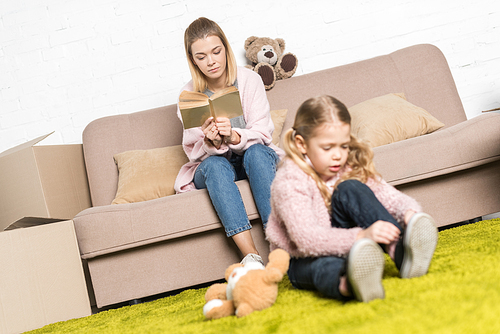 kid playing with teddy bear on carpet while mother reading book on sofa