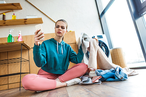 young woman using smartphone while holding clothes and sitting near washing machine