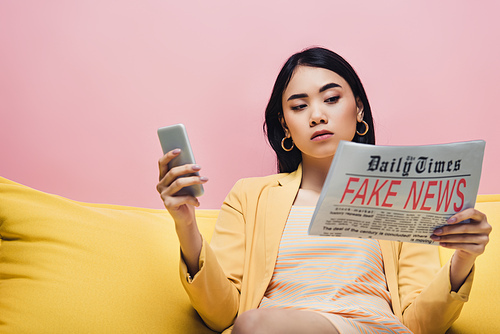 serious asian woman holding newspaper with fake news and smartphone on yellow sofa isolated on pink