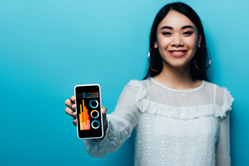 selective focus of smiling asian woman in white blouse holding smartphone with business app on blue background