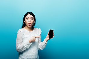 shocked asian woman pointing with finger at smartphone with blank screen on blue background