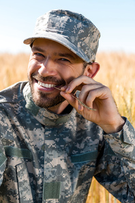 happy soldier in military uniform and cap holding wheat near lips