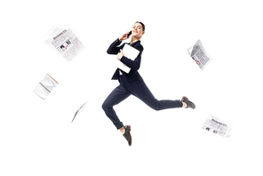 smiling businesswoman talking on smartphone and holding laptop while levitating surrounded with newspapers flying around isolated on white