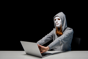 anonymous girl in mask and hoodie sitting near laptop and stretching hands during cyberbullying isolated on black