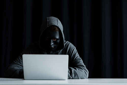 anonymous internet troll in mask typing on laptop keyboard on black