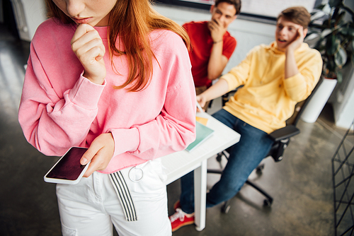 partial view of sad girl holding smartphone while classmates laughing at her in school