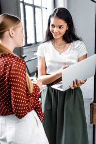 attractive multiethnic students smiling and holding laptop in apartment