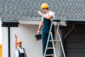 selective focus of workman waving hand and looking at coworker standing on ladder with toolbox