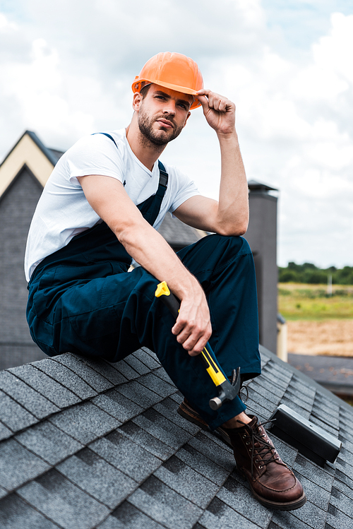 handyman in orange helmet sitting on roof and holding hammer
