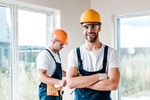 happy repairman in goggles standing with crossed arms near coworker