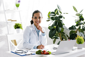 happy nutritionist in white coat near vegetables and laptop