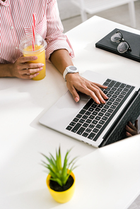 cropped view of girl using laptop while holding plastic cup with orange juice