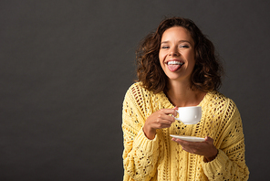 happy curly woman in yellow knitted sweater showing tongue and holding cup of coffee and saucer on black background