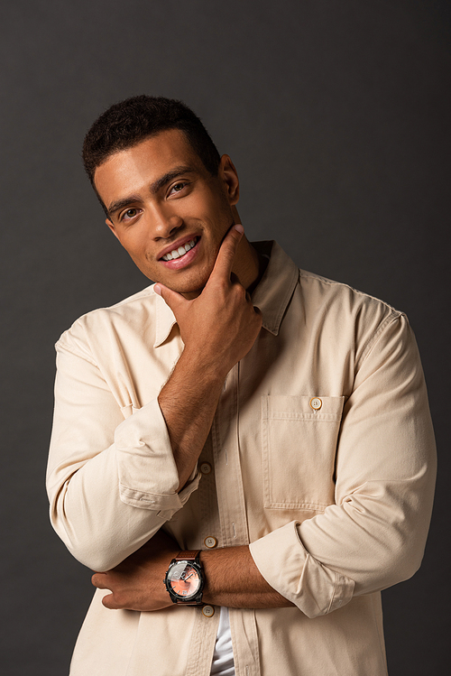 smiling handsome mixed race man in beige shirt touching face on black background