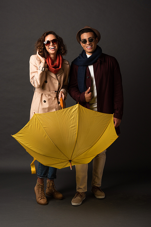 happy stylish interracial couple in autumn outfit with yellow umbrella on black background