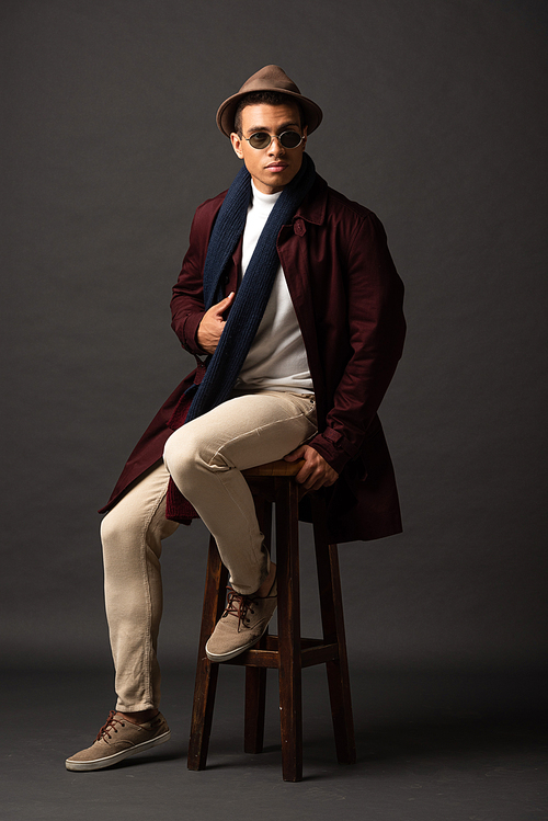 elegant mixed race man in scarf, sunglasses and hat sitting on chair on black background