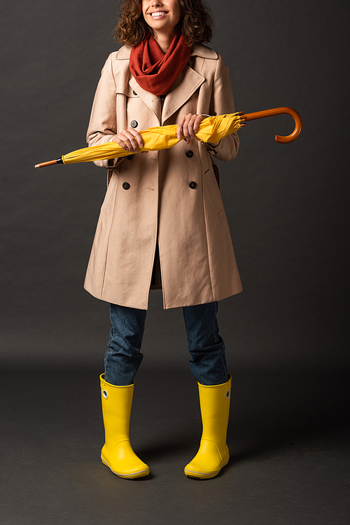 cropped view of smiling woman in trench coat and rubber boots holding yellow umbrella on black background