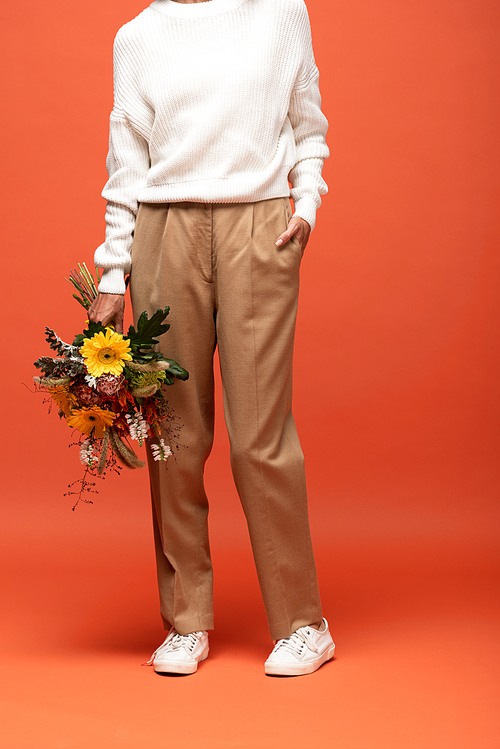 partial view of woman in autumnal outfit with hand in pocket holding bouquet of flowers on orange