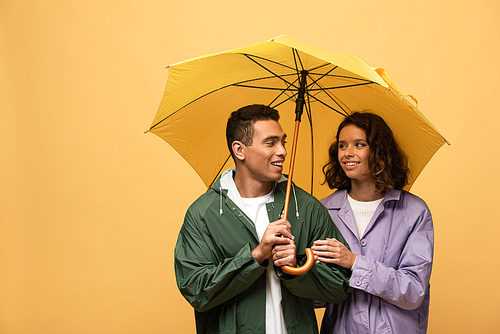 smiling interracial couple in raincoats holding umbrella isolated on yellow