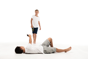 boy pretending injured while lying and aiming with toy gun at brother on white background