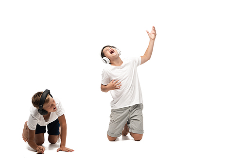 excited boy listening music in headphones and singing near laughing brother on white background