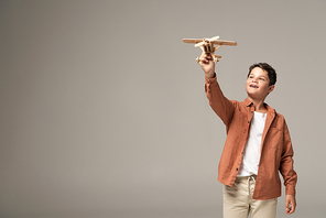 happy boy holding wooden toy plane in raised hand isolated on grey