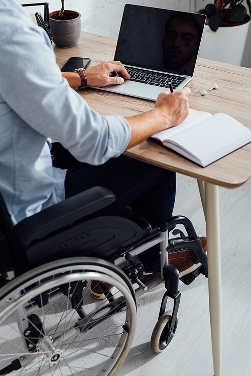 Cropped view of man in wheelchair writing in notebook and using laptop at desk