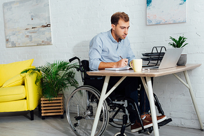 Businessman in wheelchair looking at laptop and writing in notebook at desk