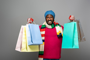 happy african american man in winter outfit with shopping bags on grey background