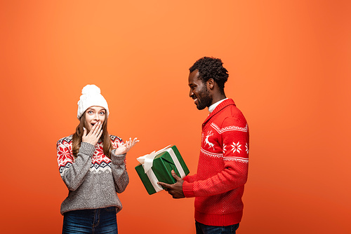 african american man gifting Christmas present to shocked girlfriend on orange background