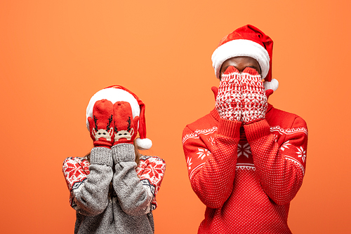 happy interracial couple in santa hats, mittens and Christmas sweaters covering face with hands on orange background