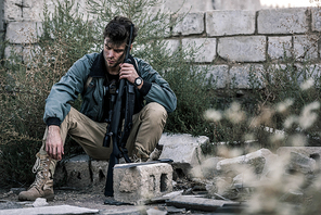 selective focus of handsome man sitting with gun near brick wall outside, post apocalyptic concept