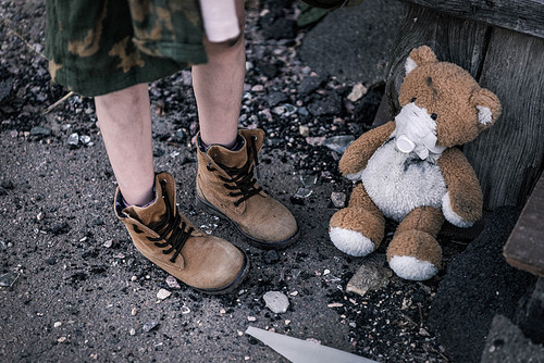 cropped view of kid in boots standing near teddy bear on ground, post apocalyptic concept