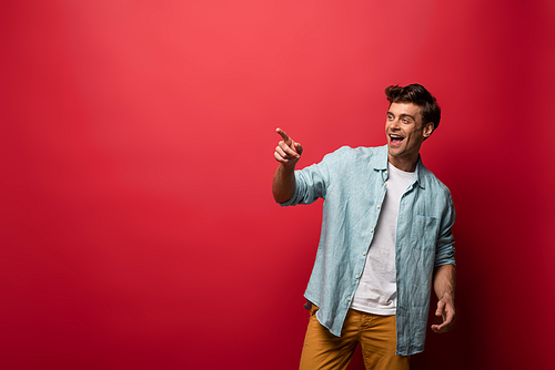 handsome laughing man in casual clothes pointing isolated on red