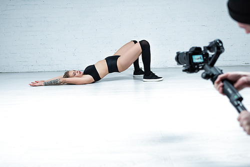 selective focus of cameraman shooting sexy blonde girl in black outfit twerking on floor near white brick wall
