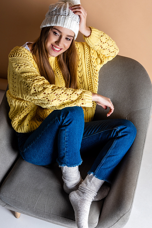 cheerful woman in yellow sweater and hat sitting in armchair on beige