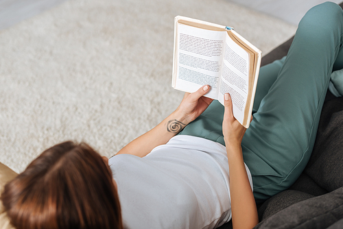 overhead view of young woman reading book while chilling on sofa in living room