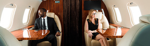 panoramic shot of handsome african american businessman and attractive businesswoman sitting in private plane