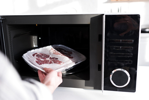 cropped view of woman putting meat into microwave in kitchen