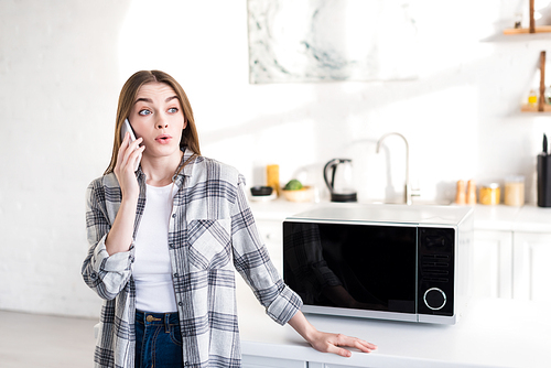 shocked woman talking on smartphone near microwave in kitchen