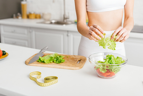 Cropped view of fit girl preparing fresh salad with avocado near measuring tape on kitchen table