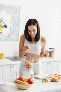 Smiling fit sportswoman adding fresh fruits in blender on kitchen table