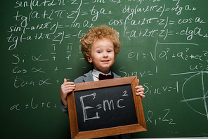 happy kid in suit and bow tie holding small blackboard with formula near chalkboard