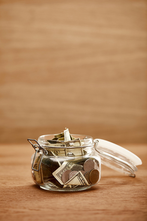 opened glass jar with dollar banknotes and coins on wooden table