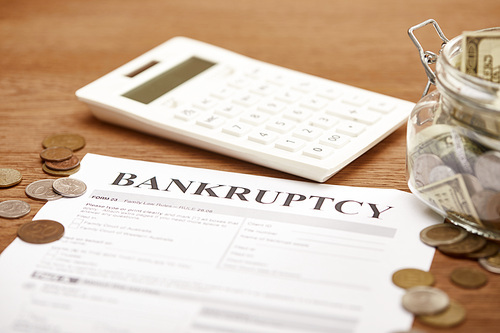 selective focus of bankruptcy form, coins, calculator and glass jar with dollar banknotes on wooden table